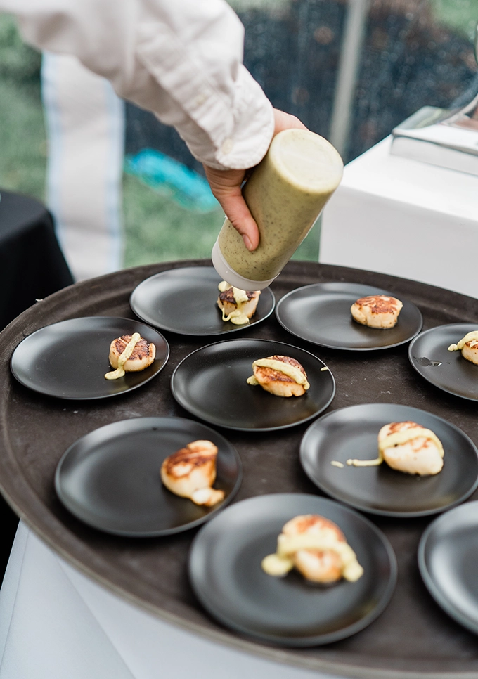 Undistinguishable person in catering attire squeezing an aioli onto a perfectly seared scallop on a black appetizer plate