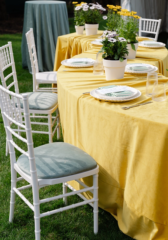 Table outfitted with a yellow table linen, white plates and white chairs with a blue cushion