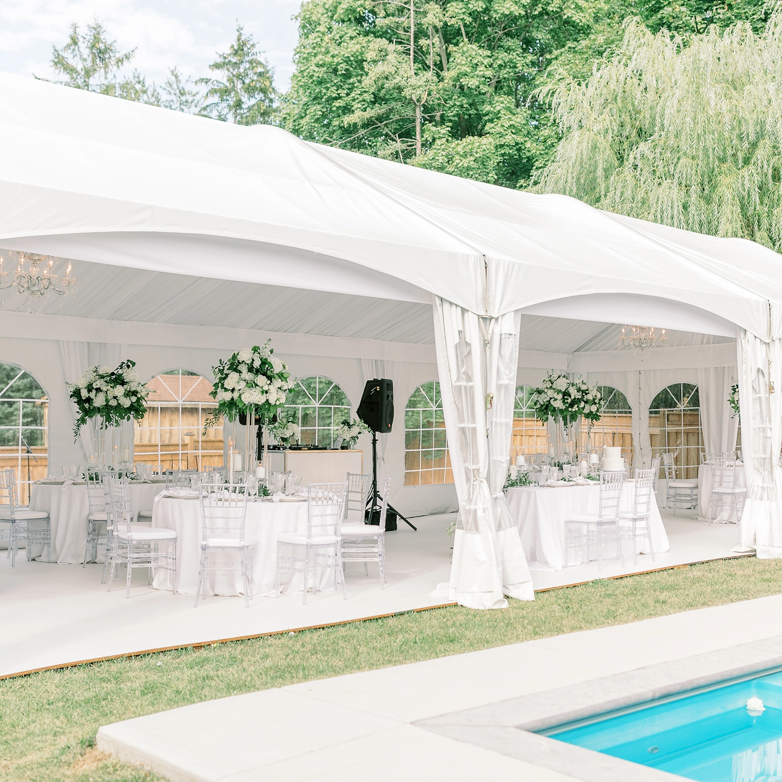 White tent rental for outdoor wedding next to in-ground pool. Tent outfitted with round tables and audio system