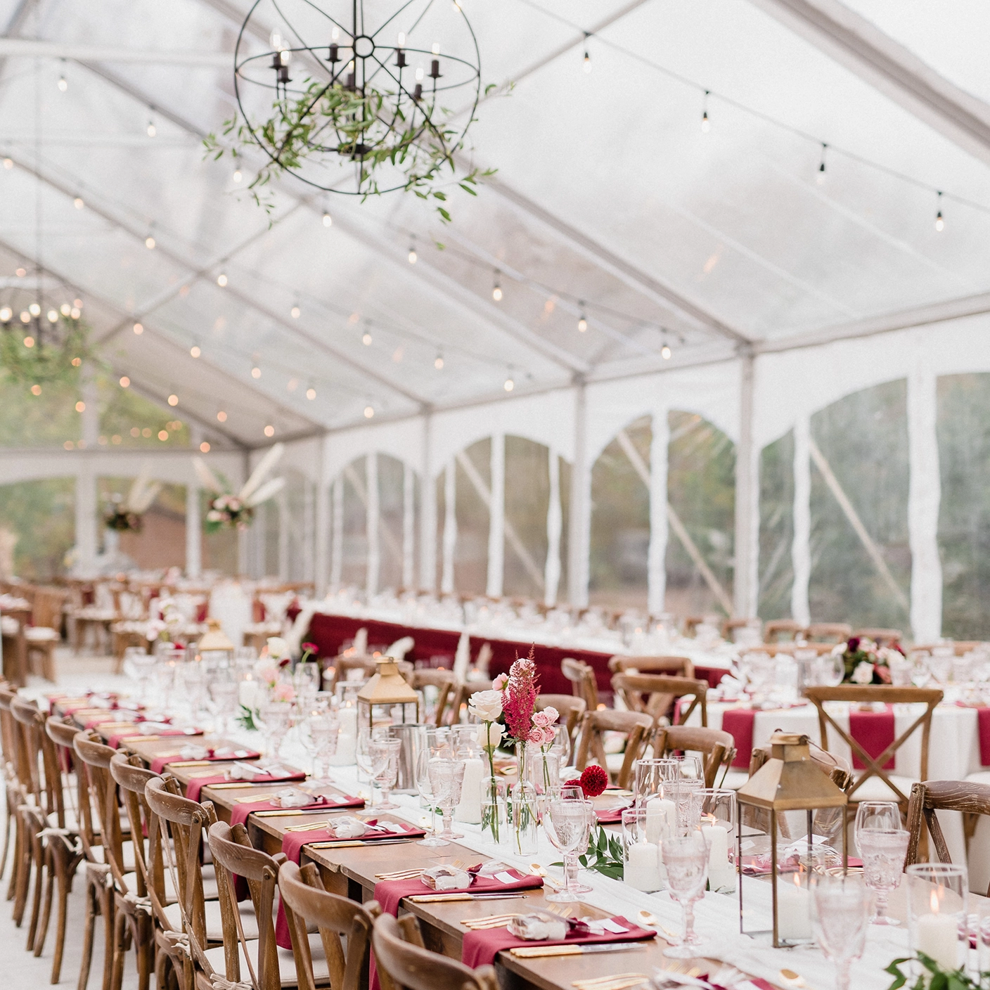 A legacy tent with clear panels and string lights strung up across the ceiling. Wooden table and chairs sit inside the tent with red and white tablescape accessories